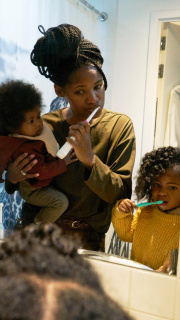 A mother holding her toddler whilst she and her other daughter brush their teeth.