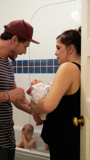 A baby having a bath and its parents talking whilst holding their other baby.